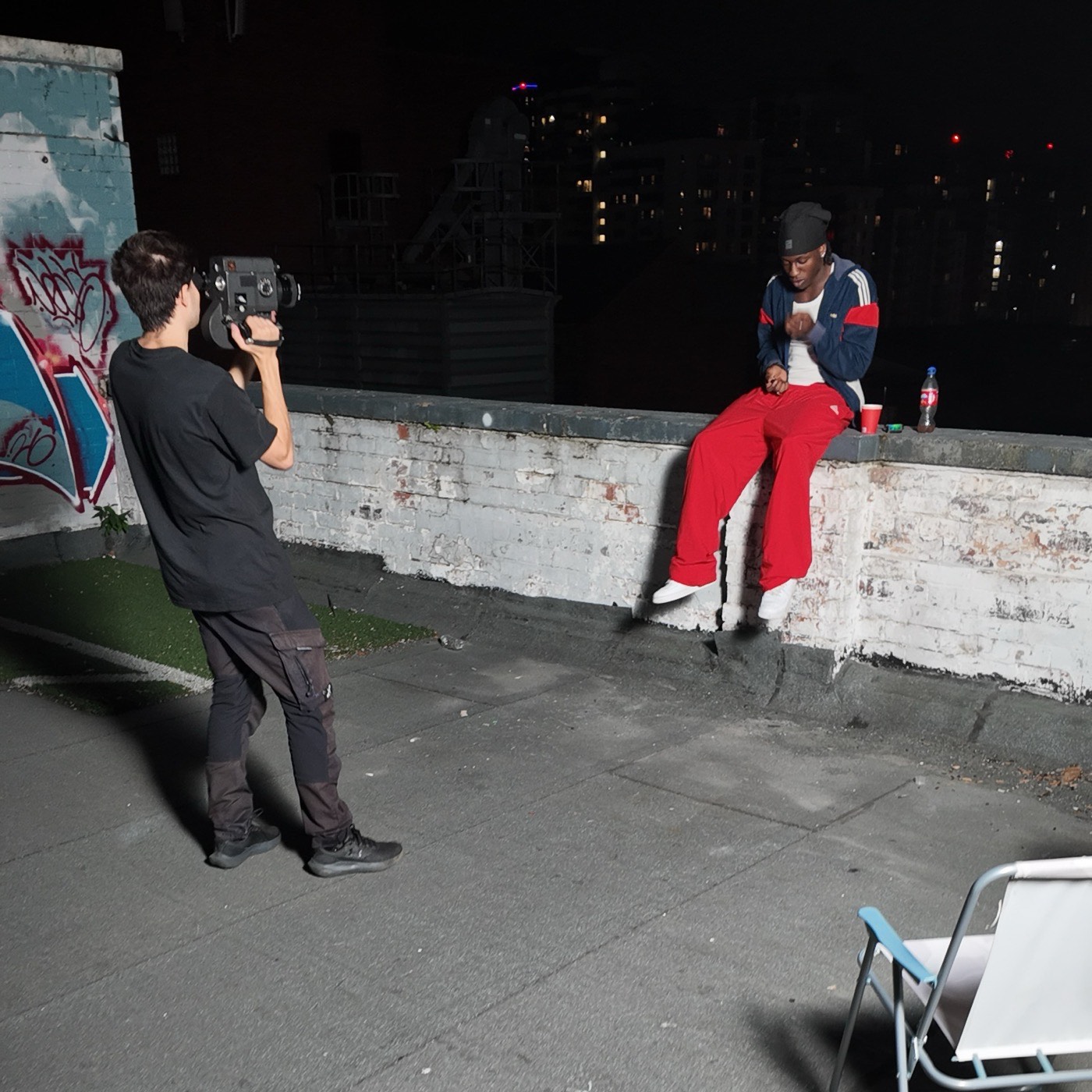 A rooftop film shoot at night — a videographer filming a musician in a red tracksuit